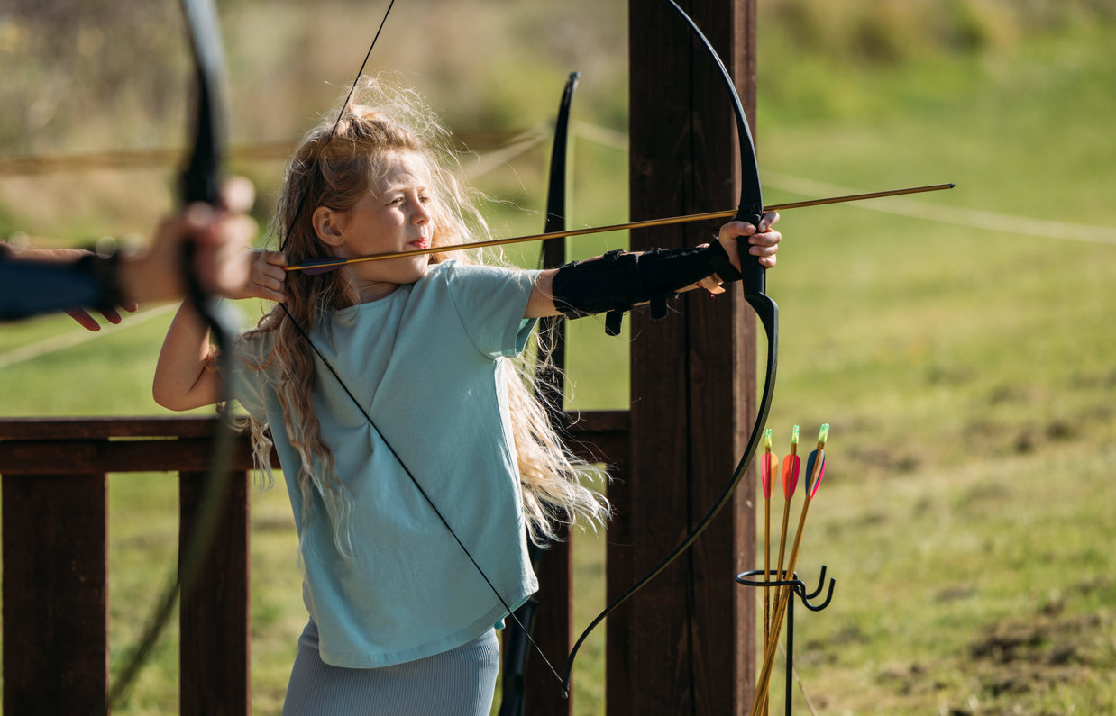 A young girl in a field aiming an archery bow
