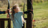 A young girl in a field aiming an archery bow