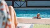 A young boy smiling in a heated outdoor pool with his arms propping him up on the side beside a woman's legs on a sun lounger on a sunny day