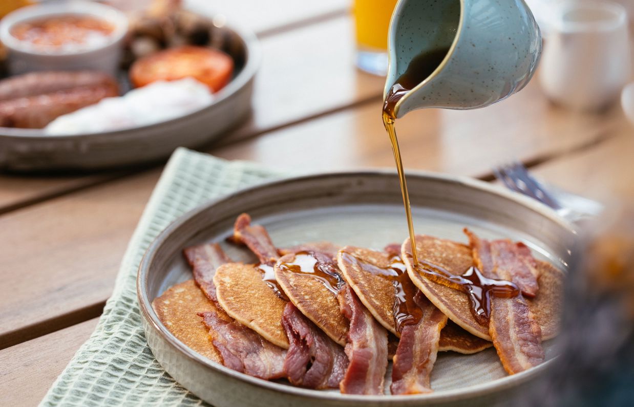 A plate of pancakes with bacon and syrup being poured over top with a full English in the background