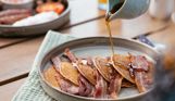 A plate of pancakes with bacon and syrup being poured over top with a full English in the background
