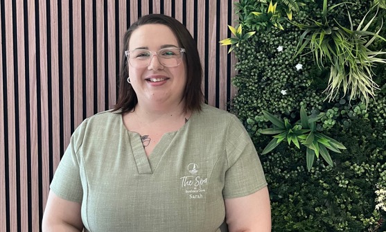 A lady smiling in spa uniform against a greenery / walnut decór background