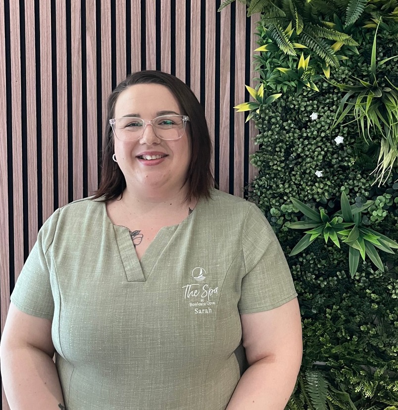 A lady smiling in spa uniform against a greenery / walnut decór background