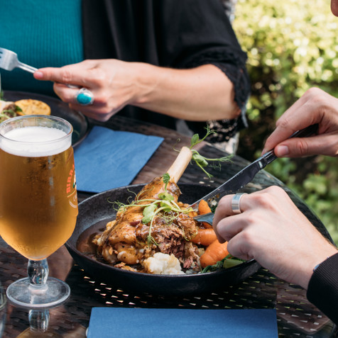 Two people sat outside eating hearty food dishes with a white wine and a pint of beer