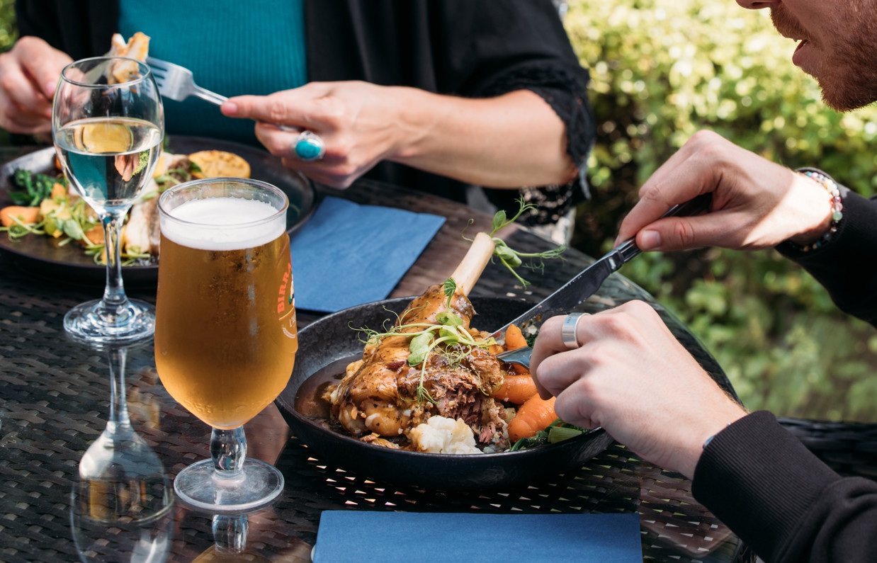 Two people sat outside eating hearty food dishes with a white wine and a pint of beer