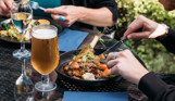 Two people sat outside eating hearty food dishes with a white wine and a pint of beer
