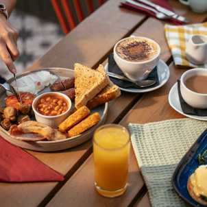 A full English breakfast and an eggs benedict on an outdoor table with various breakfast drinks and someone cutting into a tomato on the plate