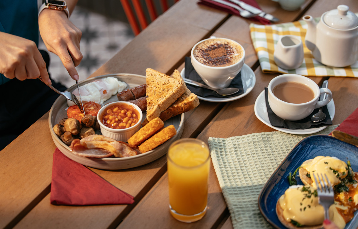 A full English breakfast and an eggs benedict on an outdoor table with various breakfast drinks and someone cutting into a tomato on the plate