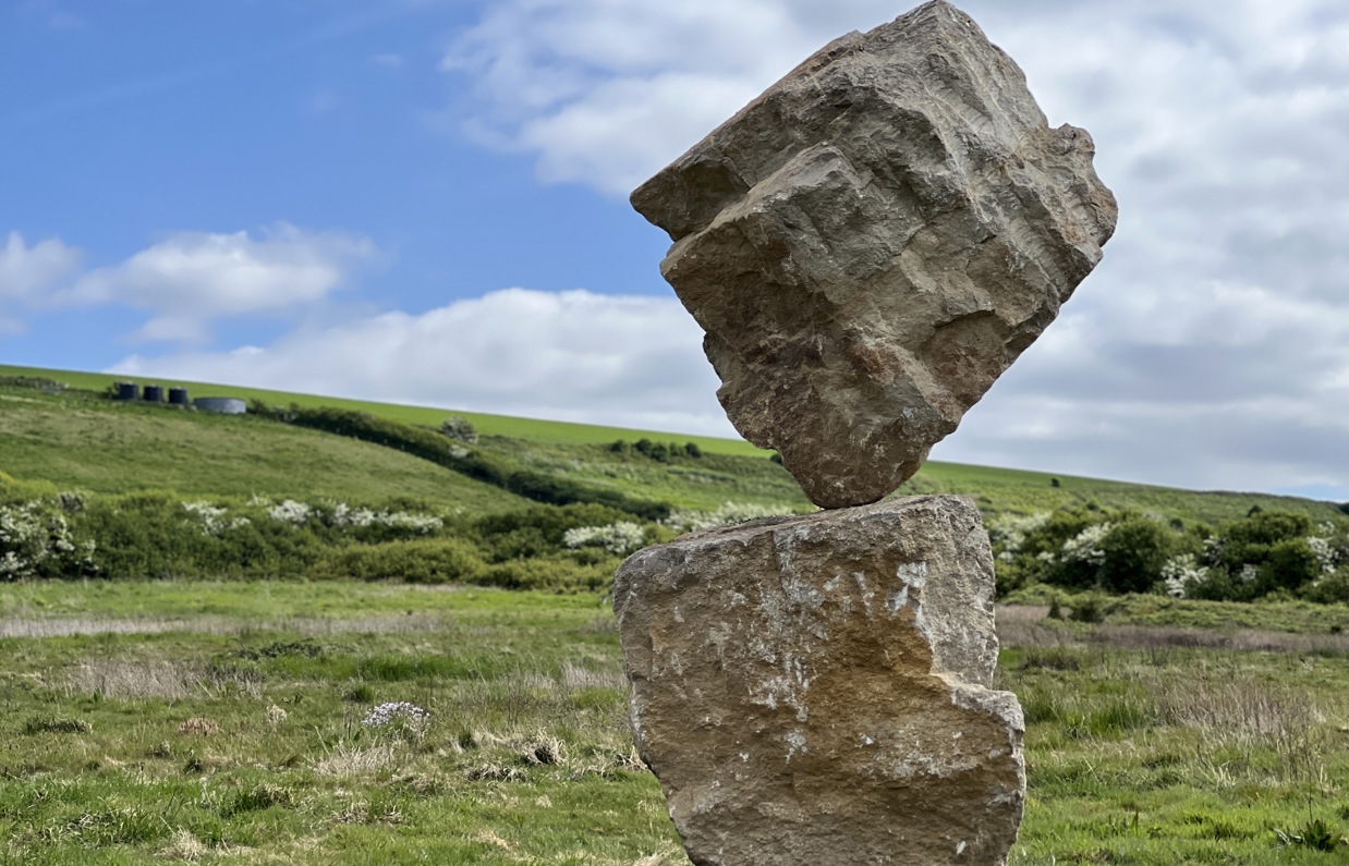 A carefully balanced stone sculpture in a countryside field with blue skies and clouds in the background