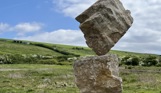 A carefully balanced stone sculpture in a countryside field with blue skies and clouds in the background