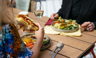 A man and a woman sat at an outside table eating a chicken sandwich and a salad with a glass of white wine and a pint of beer