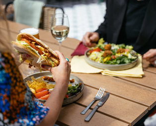 A man and a woman sat at an outside table eating a chicken sandwich and a salad with a glass of white wine and a pint of beer