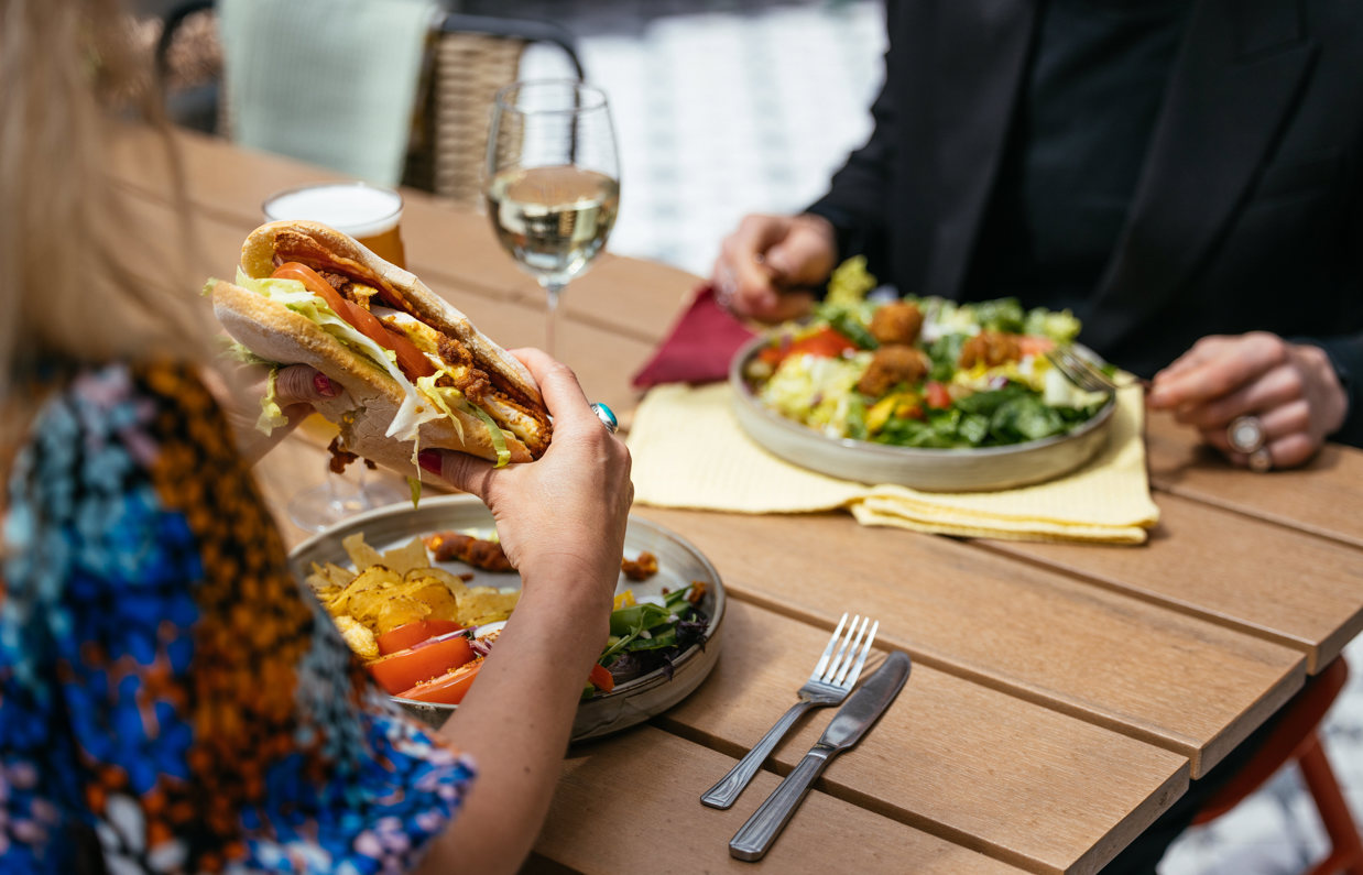 A man and a woman sat at an outside table eating a chicken sandwich and a salad with a glass of white wine and a pint of beer
