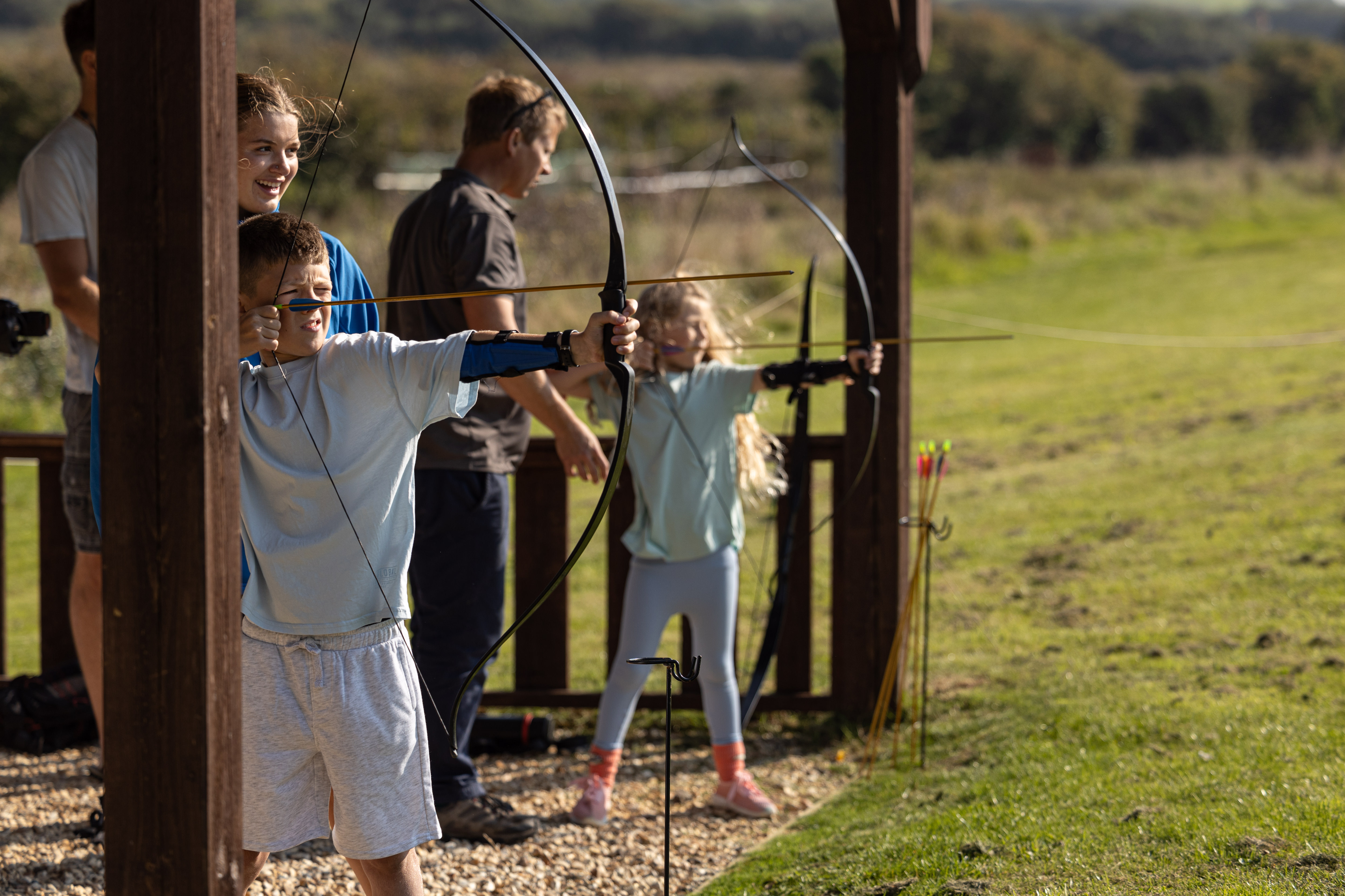 A young boy and girl taking part in archery in a dedicated activity field with adults assisting them 