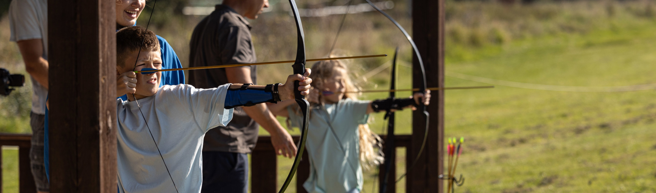 A young boy and girl taking part in archery in a dedicated activity field with adults assisting them 