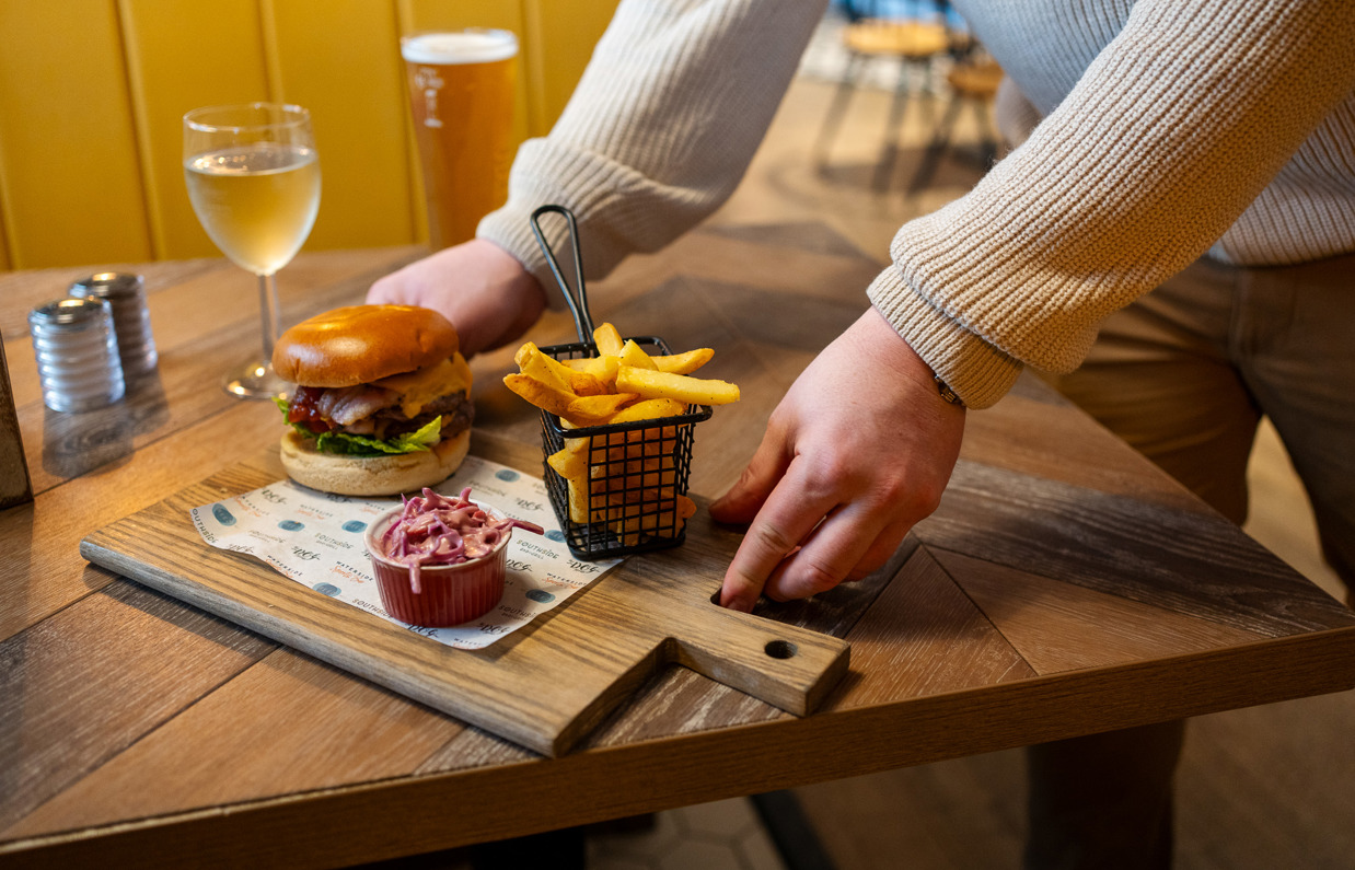 A man's hands placing a burger and chips board down on a table alongside a glass of white wine and a pint of beer