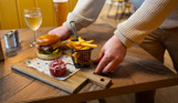 A man's hands placing a burger and chips board down on a table alongside a glass of white wine and a pint of beer