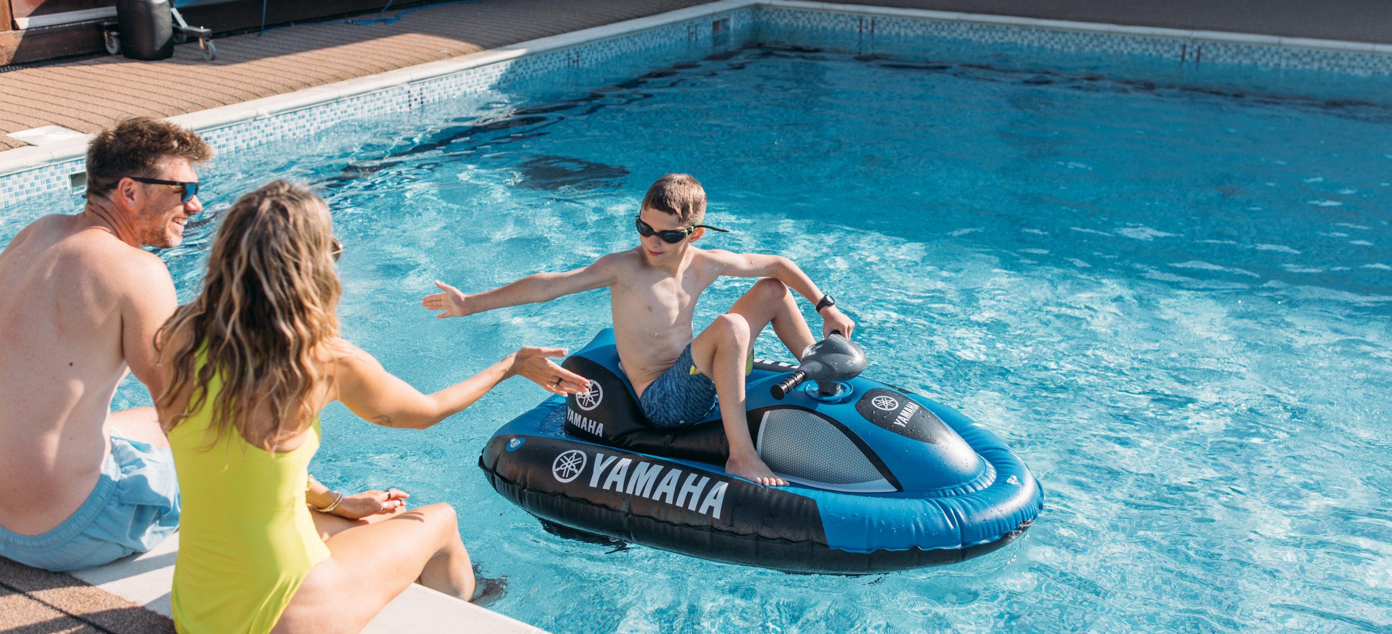 A couple sat on the edge of an outdoor swimming pool with their legs in the water whilst their son goes past on a mini jet ski high five-ing the mum