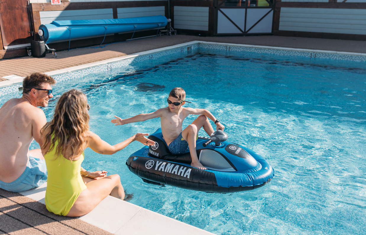 A couple sat on the edge of an outdoor swimming pool with their legs in the water whilst their son goes past on a mini jet ski high five-ing the mum