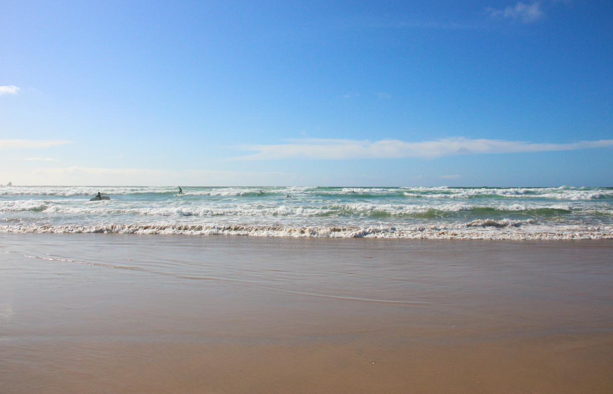 A beach with surf ready waves and people heading into surf on a sunny blue sky day