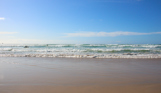 A beach with surf ready waves and people heading into surf on a sunny blue sky day