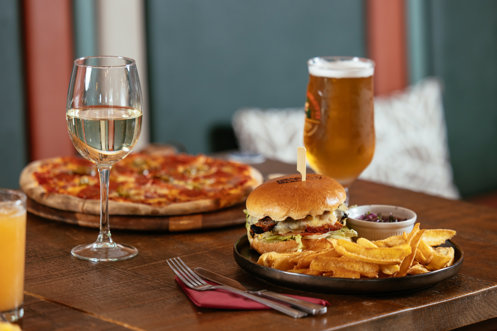 A pizza and a burger and chips on a table with a glass of white wine, a pint of beer and an orange juice