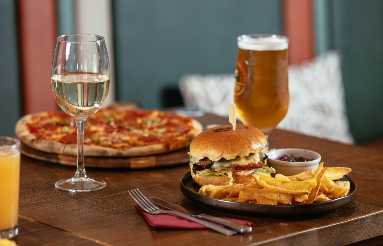 A pizza and a burger and chips on a table with a glass of white wine, a pint of beer and an orange juice