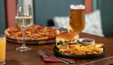 A pizza and a burger and chips on a table with a glass of white wine, a pint of beer and an orange juice
