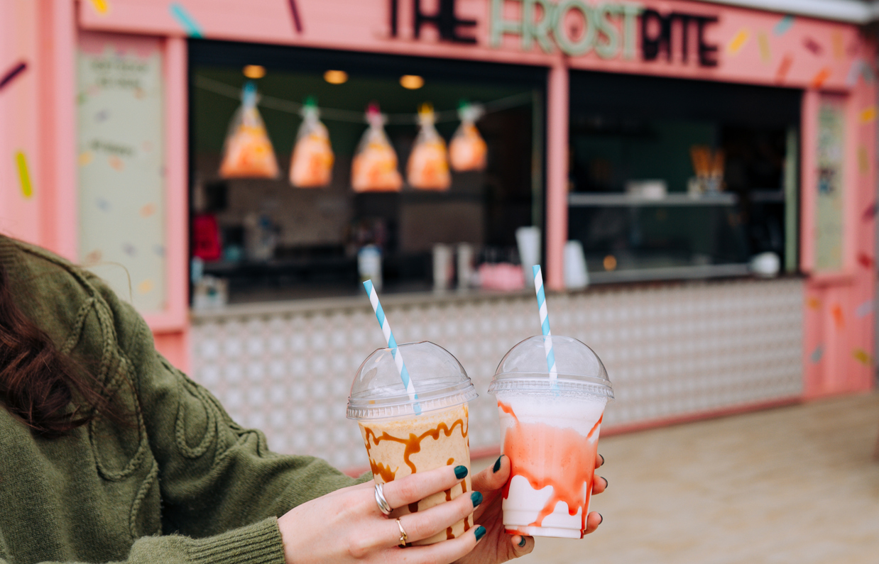Two brightly-coloured milkshakes in takeaway cups in front of a dessert bar shack