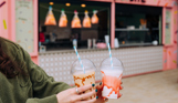 Two brightly-coloured milkshakes in takeaway cups in front of a dessert bar shack