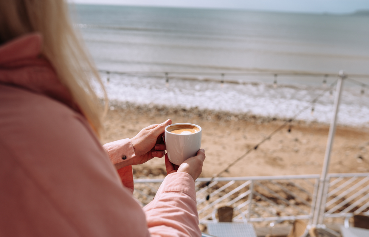 An over the shoulder shot of a woman looking out to sea from Drift Bar & Grill with a mug of coffee in her hands