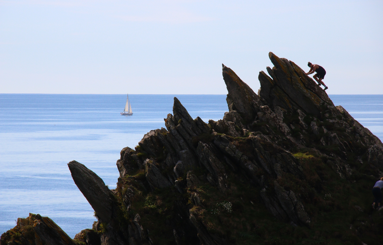 A rocky cliffside with a man climbing diagonally up it with a boat in the sea in the background