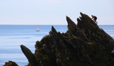 A rocky cliffside with a man climbing diagonally up it with a boat in the sea in the background