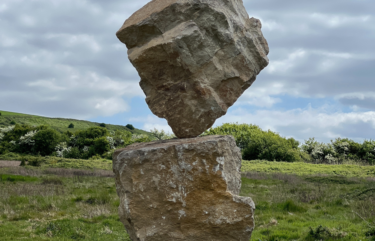 A carefully balanced stone sculpture in a countryside field with blue skies and clouds in the background