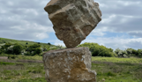 A carefully balanced stone sculpture in a countryside field with blue skies and clouds in the background