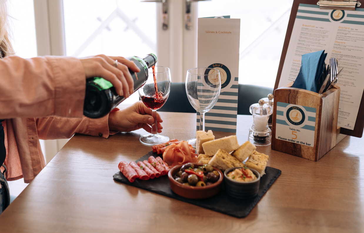 A woman sat at a table with a charcuterie board, pouring red wine into two wine glasses