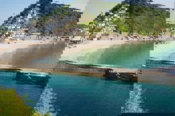 Looe beach, pier and harbour at the bottom of a green hill with blue sea waters