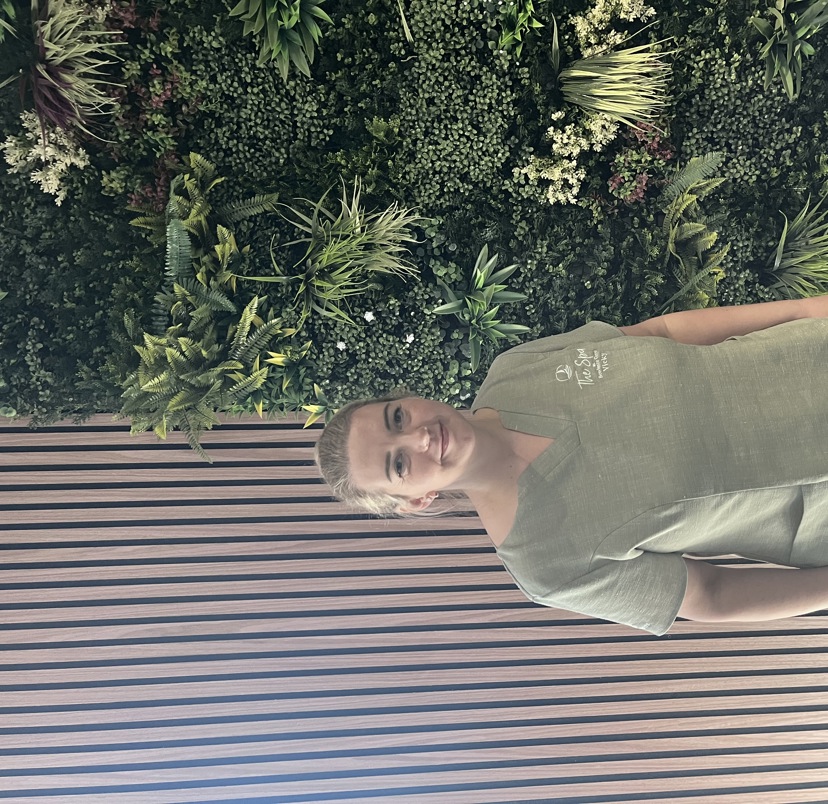 A lady smiling in spa uniform against a greenery / walnut decór background