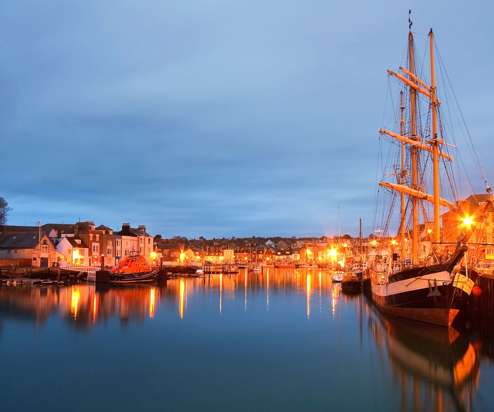 A harbour with various small boats, buildings and houses on a clear evening lit up by surrounding lights