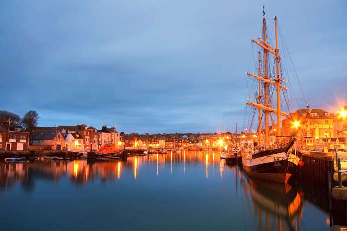 A harbour with various small boats, buildings and houses on a clear evening lit up by surrounding lights
