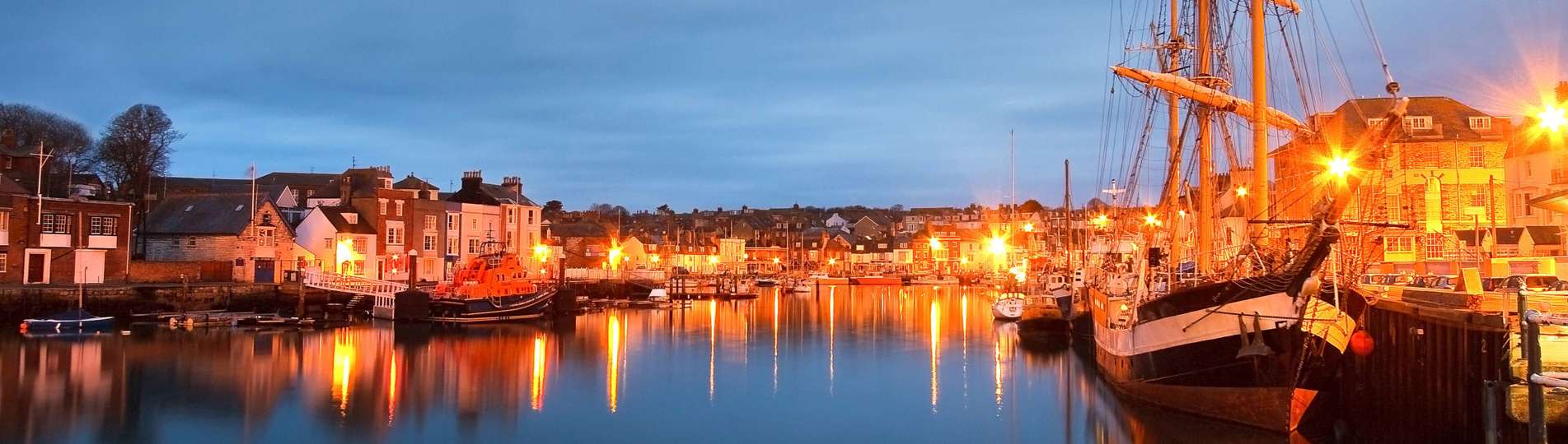 A harbour with various small boats, buildings and houses on a clear evening lit up by surrounding lights