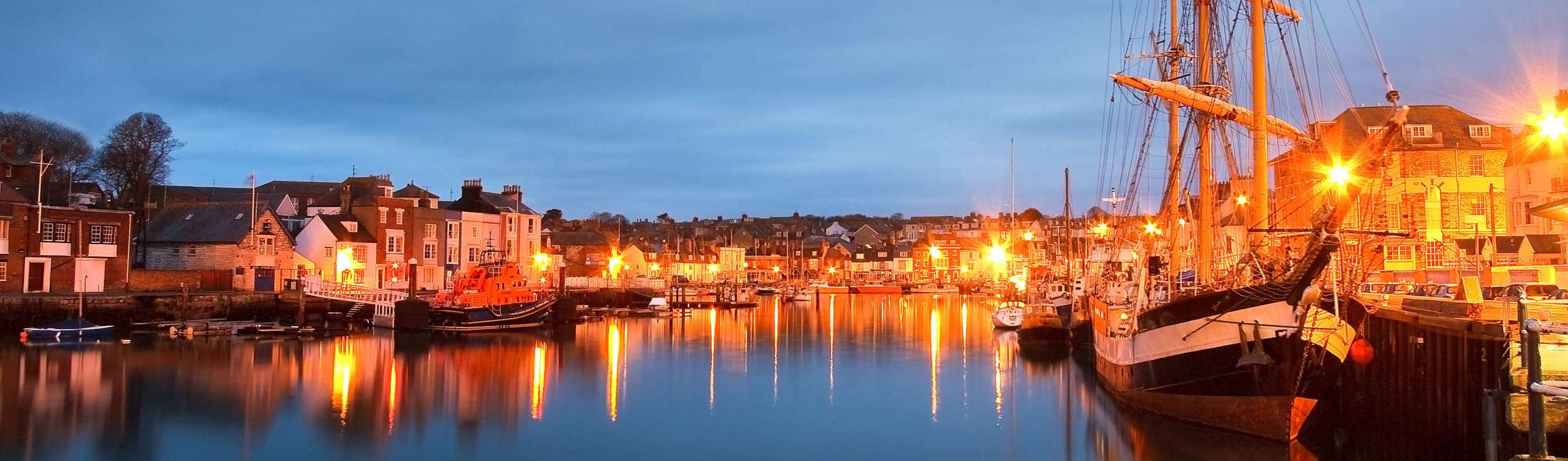 A harbour with various small boats, buildings and houses on a clear evening lit up by surrounding lights