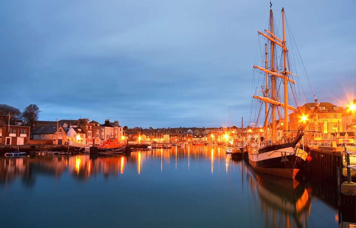 A harbour with various small boats, buildings and houses on a clear evening lit up by surrounding lights