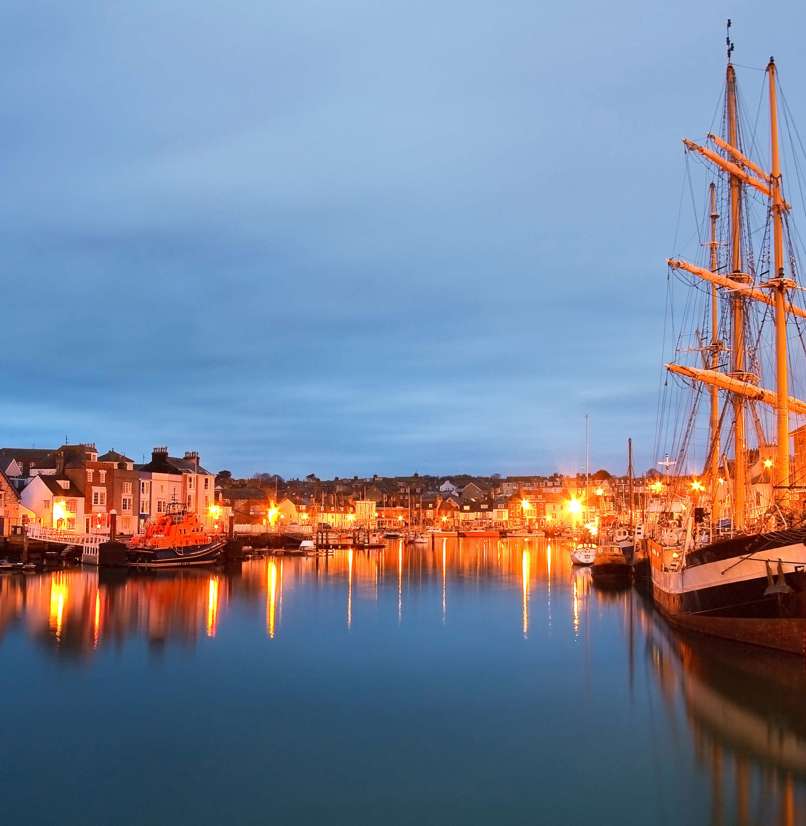 A harbour with various small boats, buildings and houses on a clear evening lit up by surrounding lights