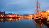 A harbour with various small boats, buildings and houses on a clear evening lit up by surrounding lights