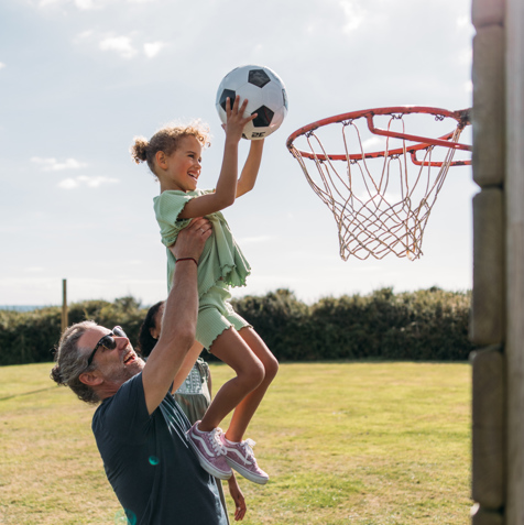 A young girl being lifted by her dad to shoot a football into a basketball hoop in a sports field with seaviews in the background