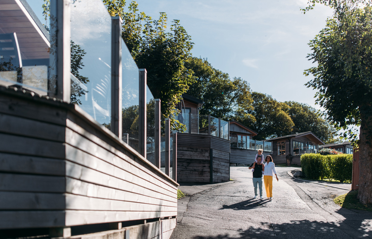 A family of three walking down a road on a lodge park surrounded by holiday lodges and woodland on a sunny, blue sky day
