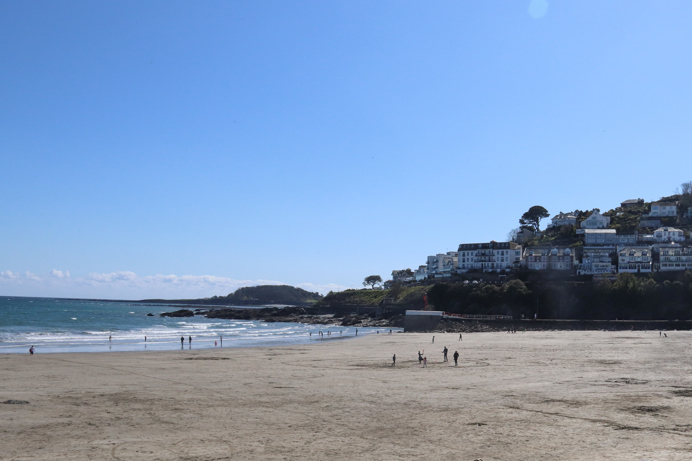 A wide stretch of beach at the bottom of a hill with houses on it on a sunny, blue sky day