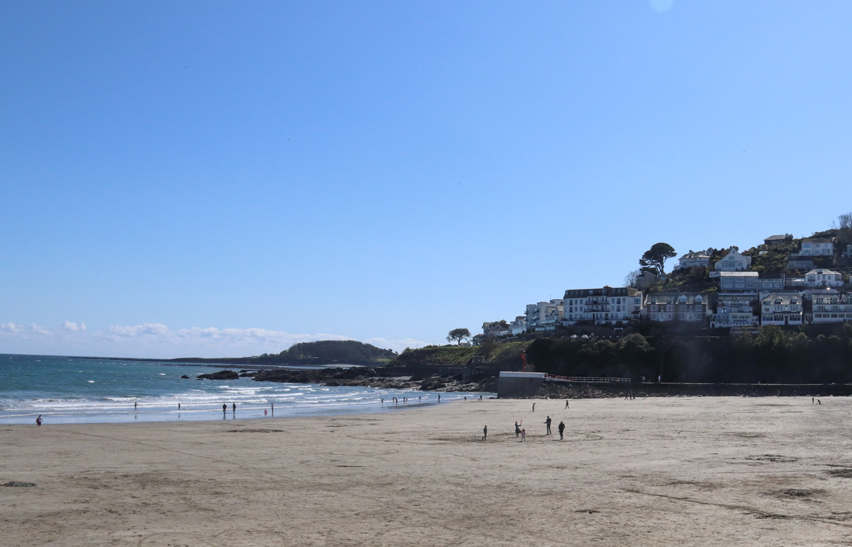 A wide stretch of beach at the bottom of a hill with houses on it on a sunny, blue sky day