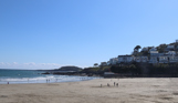 A wide stretch of beach at the bottom of a hill with houses on it on a sunny, blue sky day